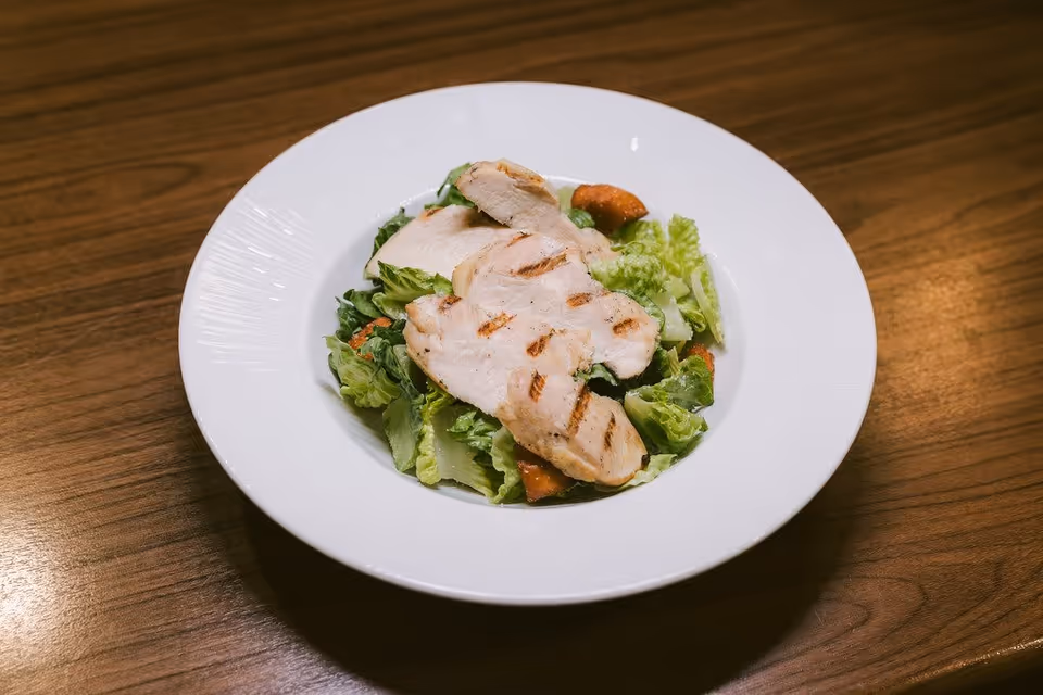 A white plate with grilled chicken breast slices served on a bed of fresh green lettuce and croutons, placed on a wooden table.