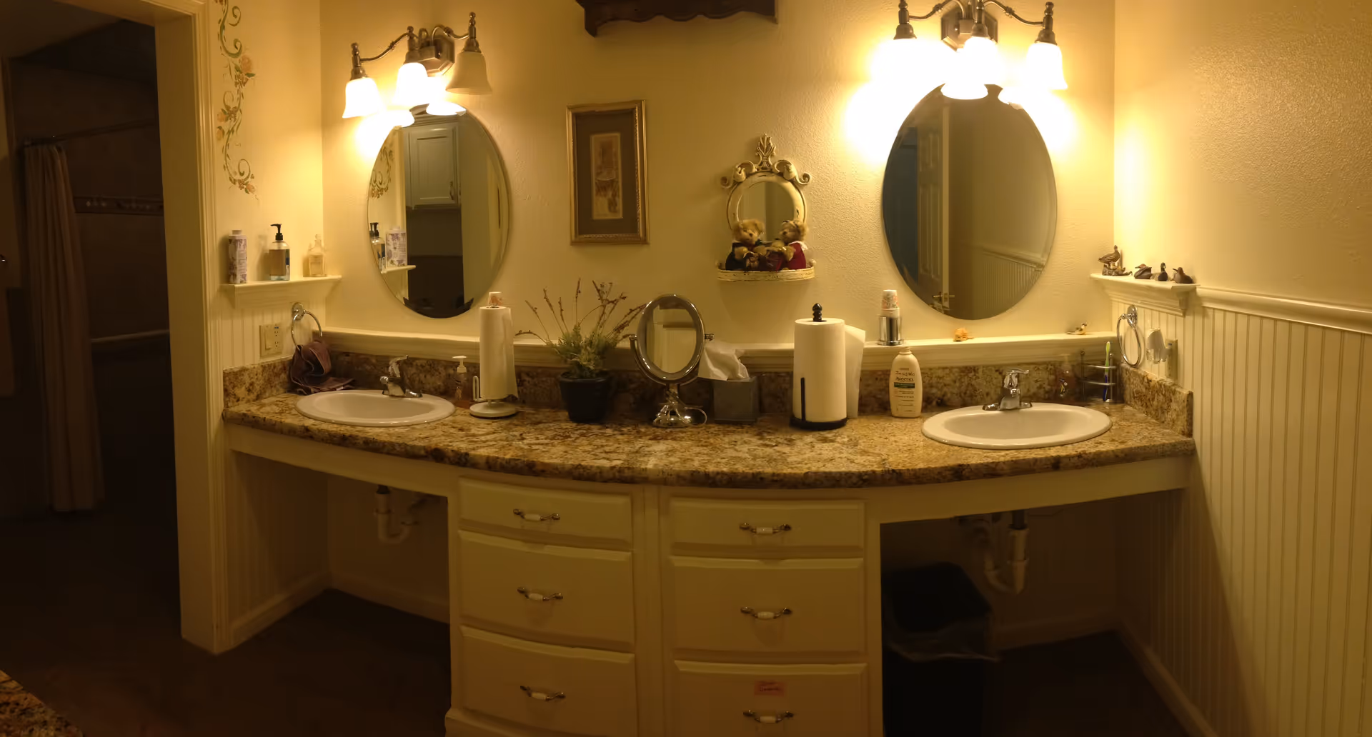 A bathroom vanity with two sinks, each under an oval mirror with three-light fixtures above. The countertop is granite with various items including soap dispensers, paper towels, a small potted plant, and a tissue box. There are decorative elements on the wall including a framed picture and a small shelf with teddy bears. The bathroom has cream-colored walls and beadboard wainscoting.