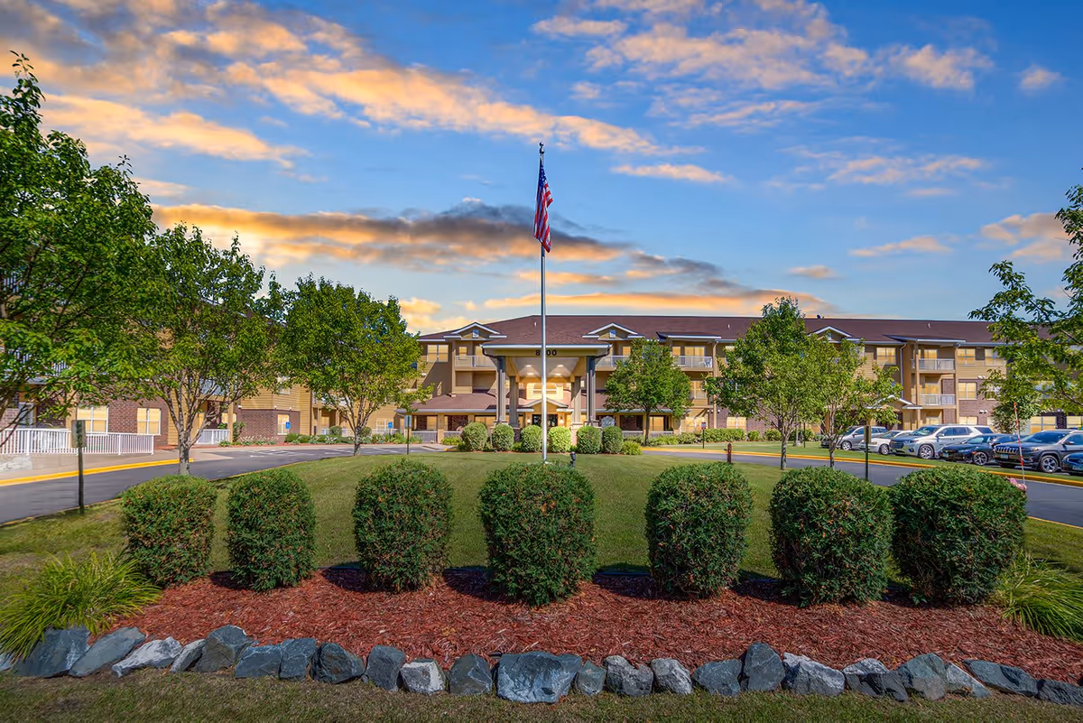 Exterior view of Tradition Living facility showing a large building with multiple floors, a central entrance with a covered porch, an American flag on a flagpole in front, surrounded by neatly trimmed bushes, trees, and a parking area with cars under a partly cloudy sky during sunset.
