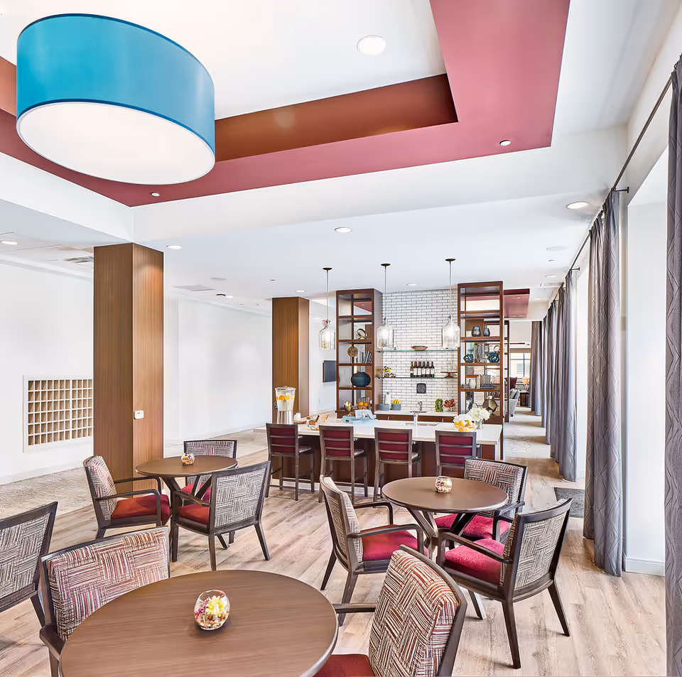 A bright and modern dining area in a senior living facility featuring round wooden tables with patterned cushioned chairs. The room has wood flooring, large windows with gray curtains, and a ceiling with a recessed maroon section and a large blue circular light fixture. In the background, there is a kitchen or bar area with high chairs, pendant lights, and open shelving displaying decorative items and bottles.