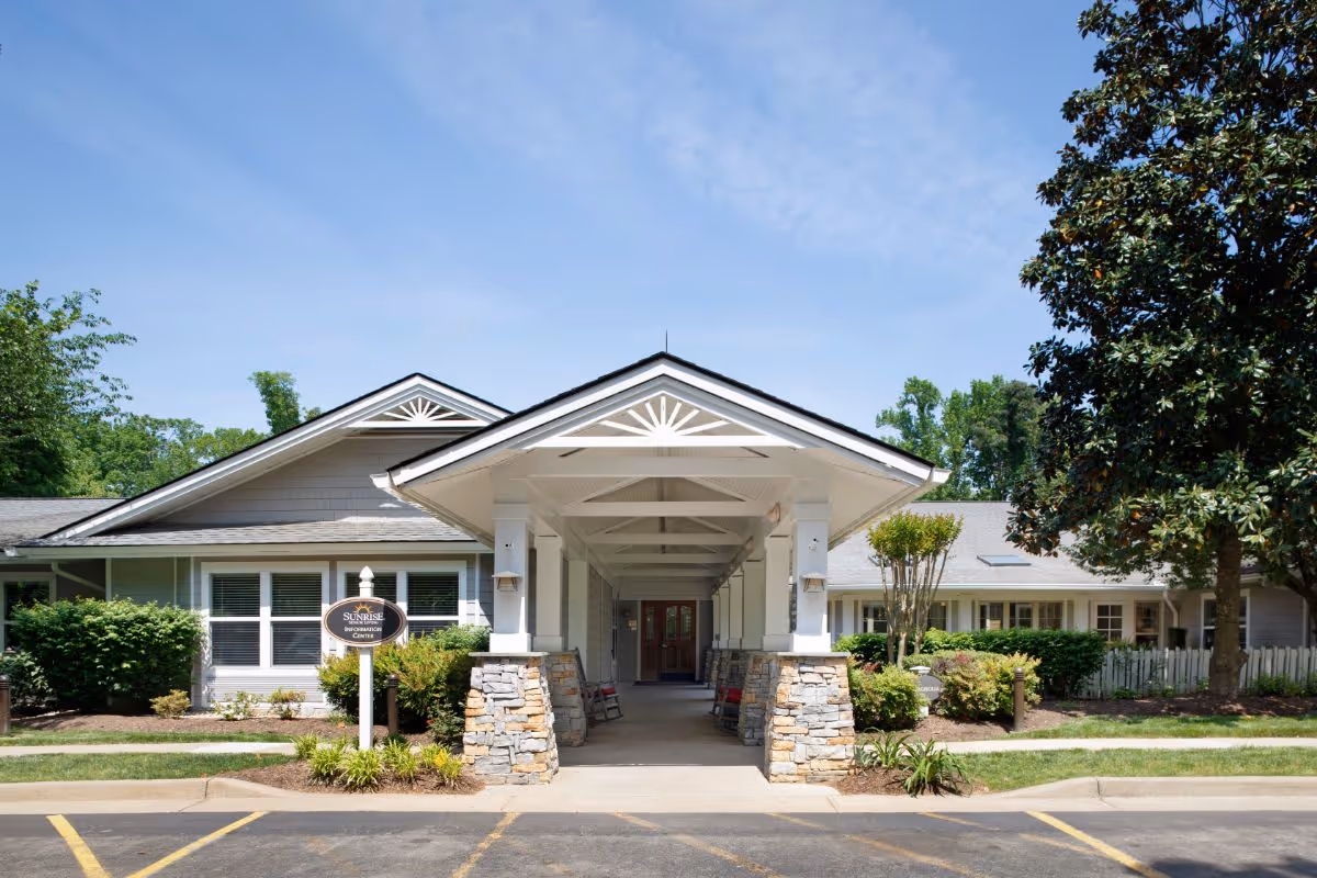 Front exterior view of Sunrise of Richmond facility with a covered entrance supported by stone pillars, surrounded by greenery and trees under a clear blue sky.