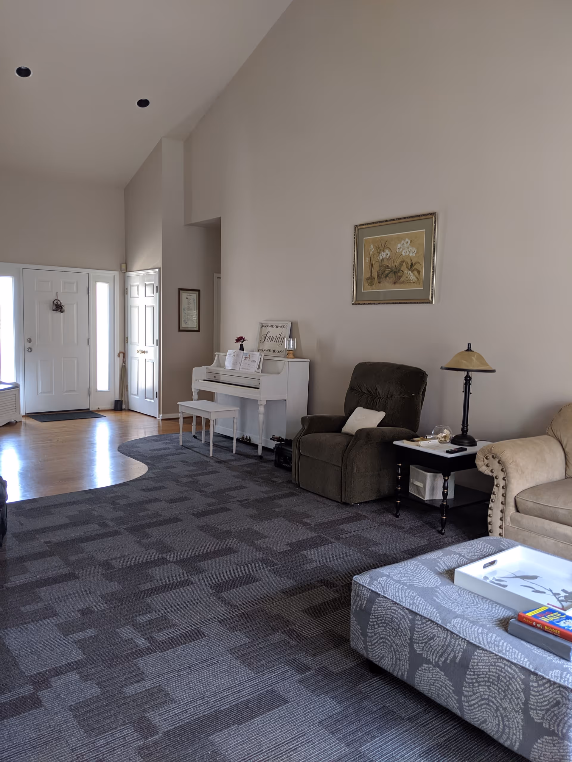 A spacious living room with a high ceiling, featuring a white upright piano with a bench, a dark recliner chair with a pillow, a side table with a lamp, a beige couch, and a patterned ottoman with a white tray and books on it. The room has a mix of carpet and hardwood flooring, and a front door with sidelights is visible in the background.