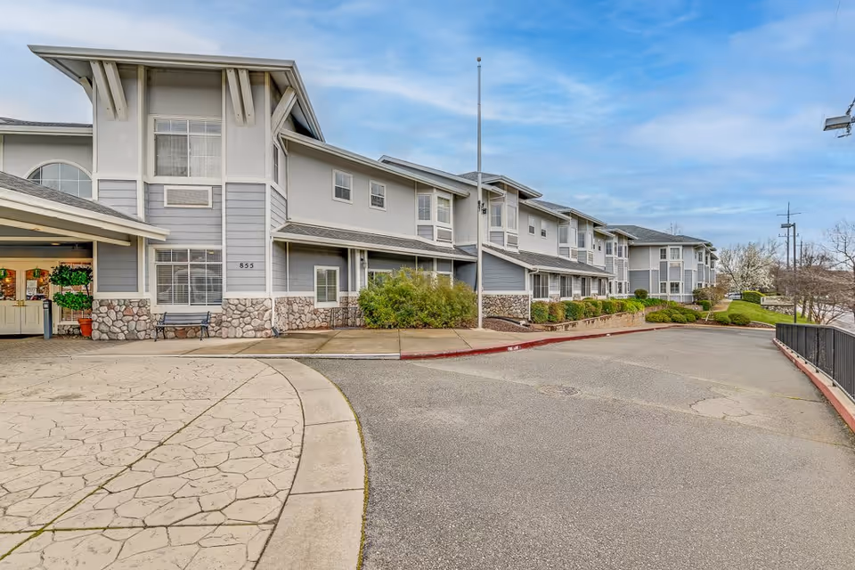 Exterior view of a senior living facility building with gray and white siding, stone accents, and multiple windows. There is a driveway and a sidewalk leading to the entrance, with some bushes and landscaping along the building. The sky is partly cloudy.
