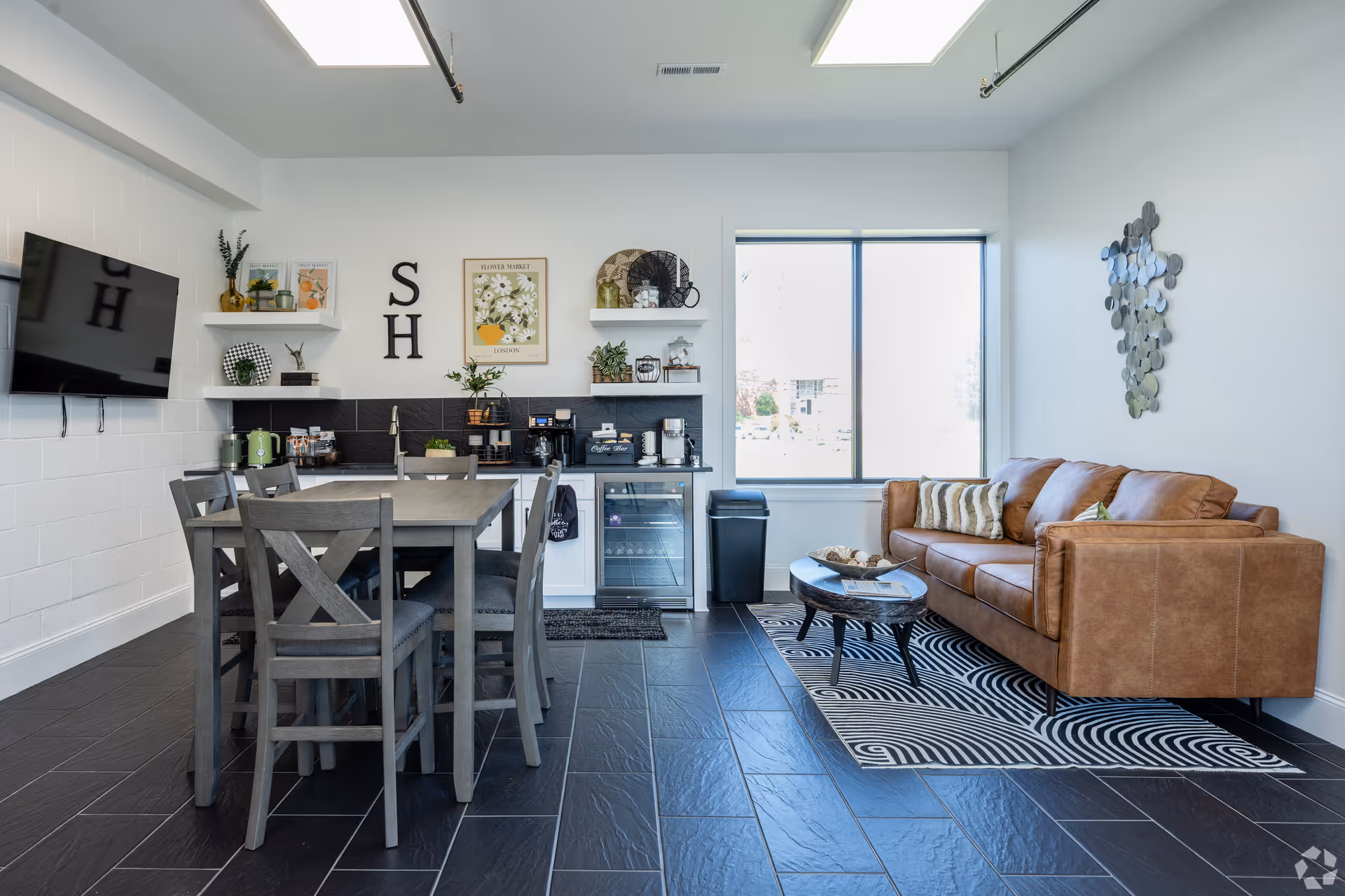 A modern common area featuring a brown leather sofa with patterned pillows on a black and white rug, a round coffee table, a dining table with six chairs, a wall-mounted TV, and shelves with decorative items and plants. There is a large window letting in natural light and a small kitchenette area with a coffee machine and mini fridge.