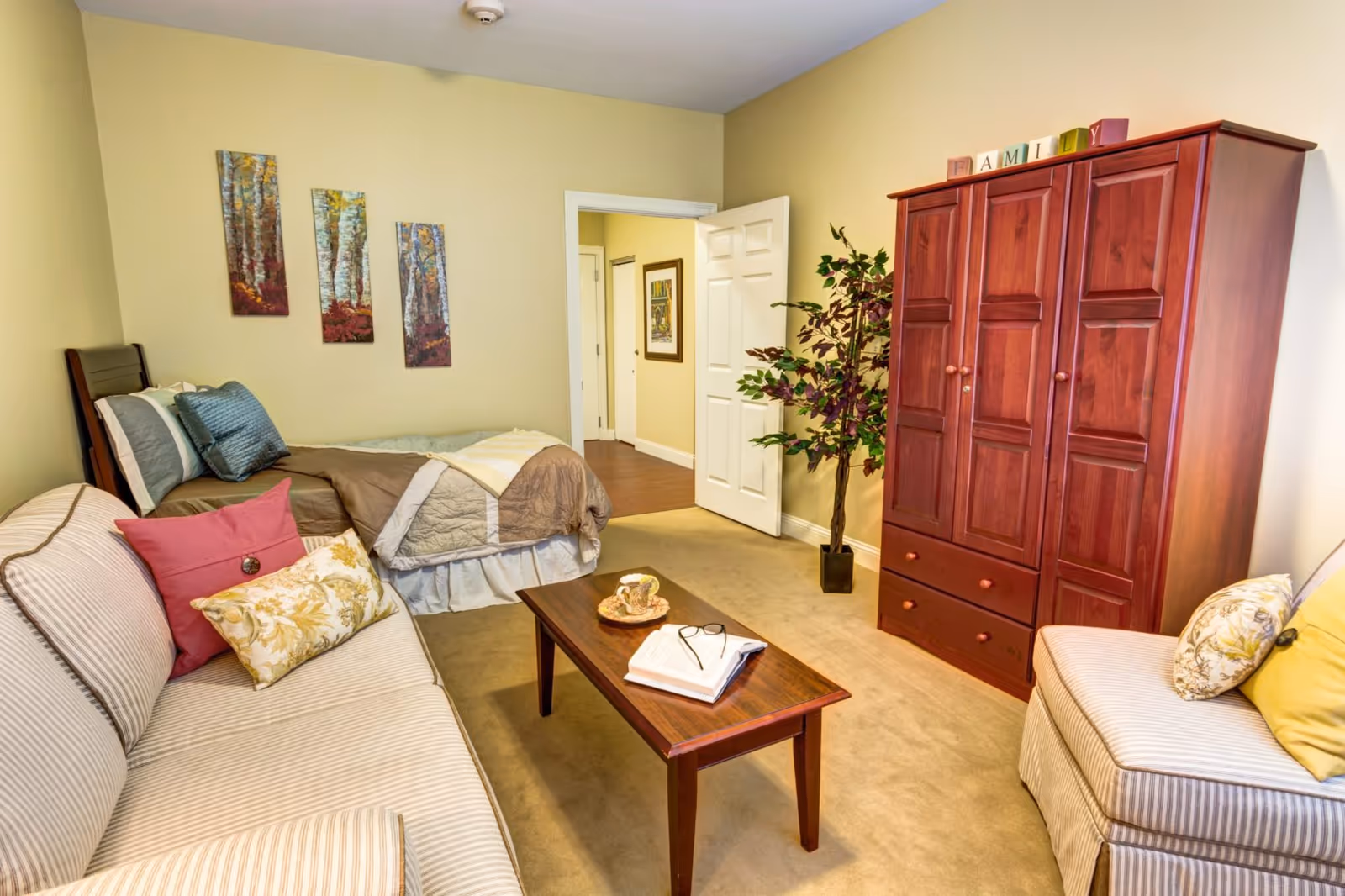 A cozy studio-style living room with a bed, striped sofa, wooden armoire, coffee table and potted plant.