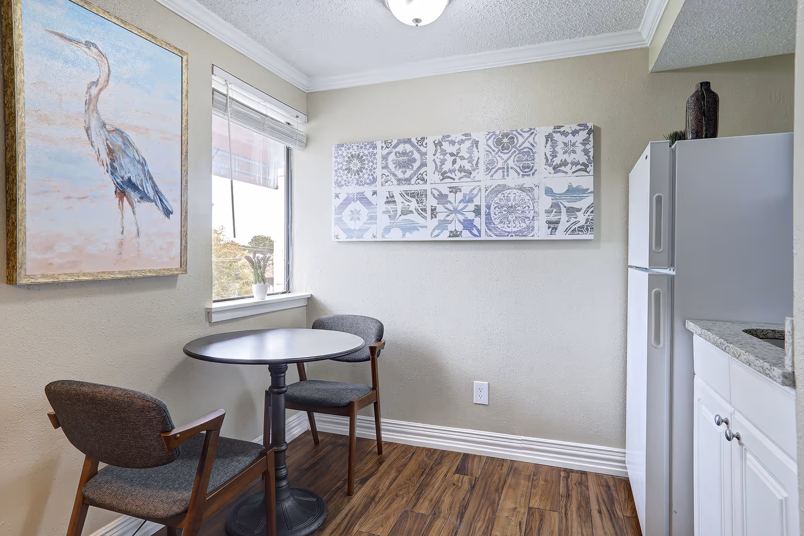 Small dining area with a round black table and two gray cushioned chairs with wooden arms. There is a window with white blinds letting in natural light. On the wall to the left, there is a framed painting of a heron bird. On the wall in front, there is a decorative artwork featuring various blue and white tile patterns. To the right, part of a white refrigerator and white cabinetry with a granite countertop are visible. The floor is wood with a warm tone.