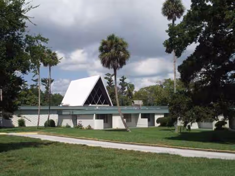 Exterior view of a low, mid-century building with a triangular white roof and palm trees.