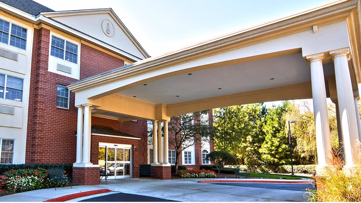 Entrance of a senior living facility with a covered drop-off area supported by white columns, red brick and beige exterior walls, surrounded by landscaped greenery and flowers.