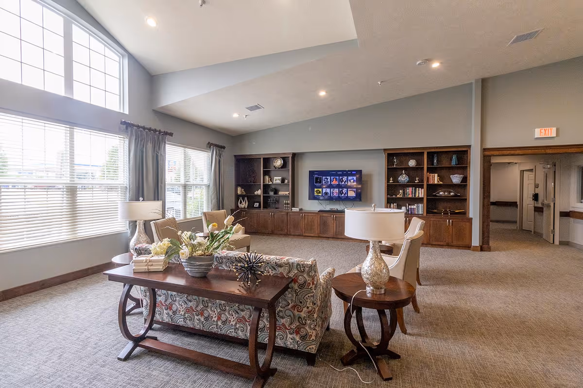 A spacious living room area in an assisted living facility with large windows letting in natural light. The room features a patterned sofa, two armchairs, a wooden coffee table with a flower arrangement, side tables with lamps, and built-in wooden shelves with decorative items and books. A flat-screen TV is mounted on the wall between the shelves.