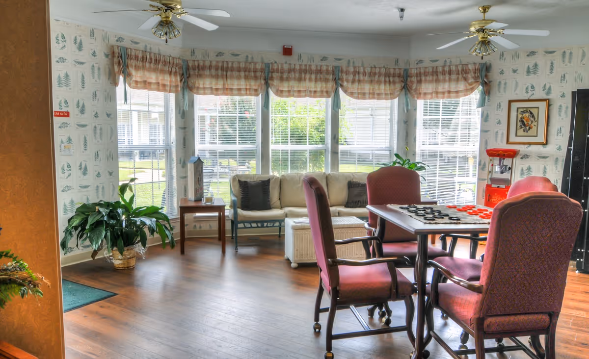 A cozy common area with large windows covered by plaid valances, letting in natural light. The room features a white cushioned bench with dark pillows, a wicker storage ottoman, a wooden side table with a decorative item, and several burgundy upholstered chairs around a table with a checkers game set up. There are green plants in pots and wallpaper with a leaf pattern on the walls. The floor is wooden, and two ceiling fans with lights are visible.