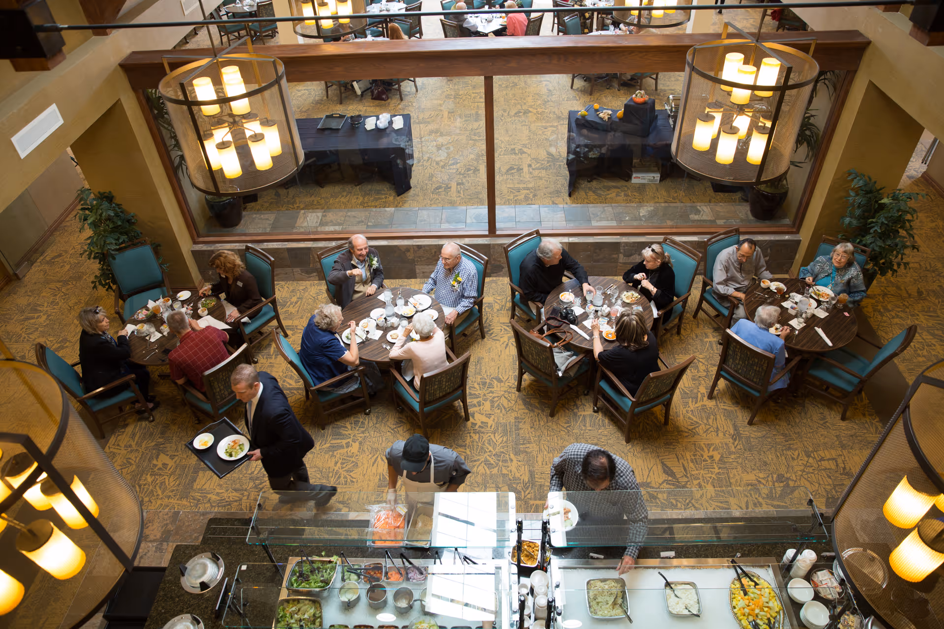 Overhead view of a retirement community dining room with seniors seated at round tables and a buffet station in the foreground.