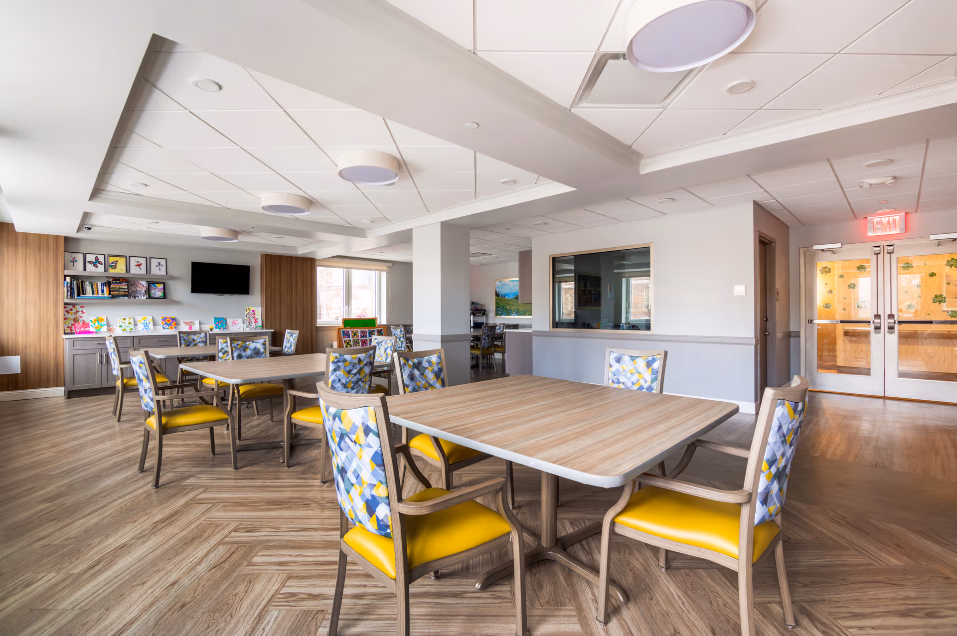 A bright and spacious common area in an assisted living facility featuring several wooden tables surrounded by chairs with yellow seats and patterned backs. The room has wood-patterned flooring, a white ceiling with recessed lighting, and large windows letting in natural light. There are bookshelves with books and colorful artwork displayed on a cabinet against the far wall, and a flat-screen TV mounted above. Double doors with glass panels and an exit sign are visible in the background.