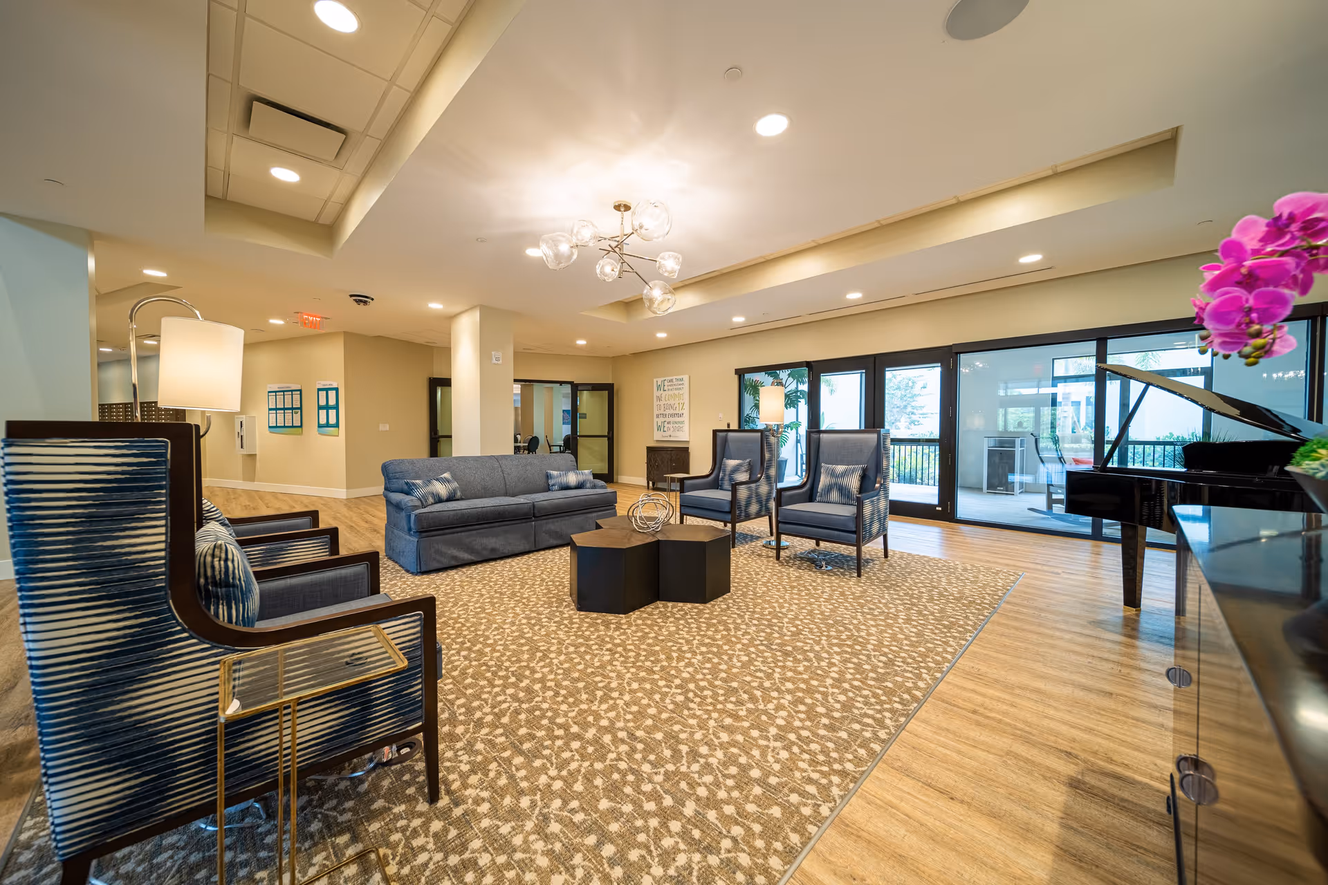 A spacious and well-lit living room area with a patterned carpet, a blue sofa, two blue armchairs, a modern chandelier, a floor lamp, and a black grand piano near large glass doors leading to an outdoor patio. There is a pink orchid flower in the foreground on the right side.