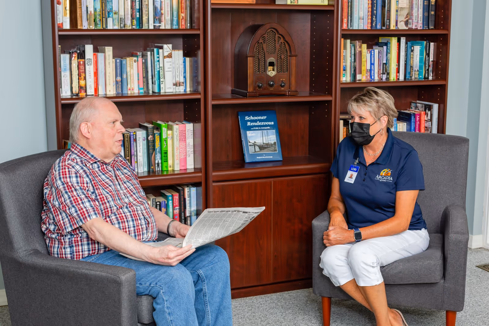 An elderly man holding a newspaper sits in a gray armchair facing a woman wearing a black face mask, a navy blue Arcadia polo shirt, and white pants, who is seated in another gray armchair. Behind them is a wooden bookshelf filled with books and a vintage radio.