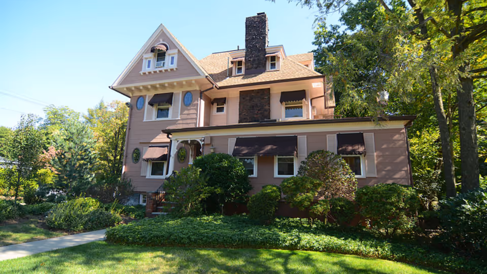 Pink multi-story house with a stone chimney, window awnings and a landscaped lawn and trees.