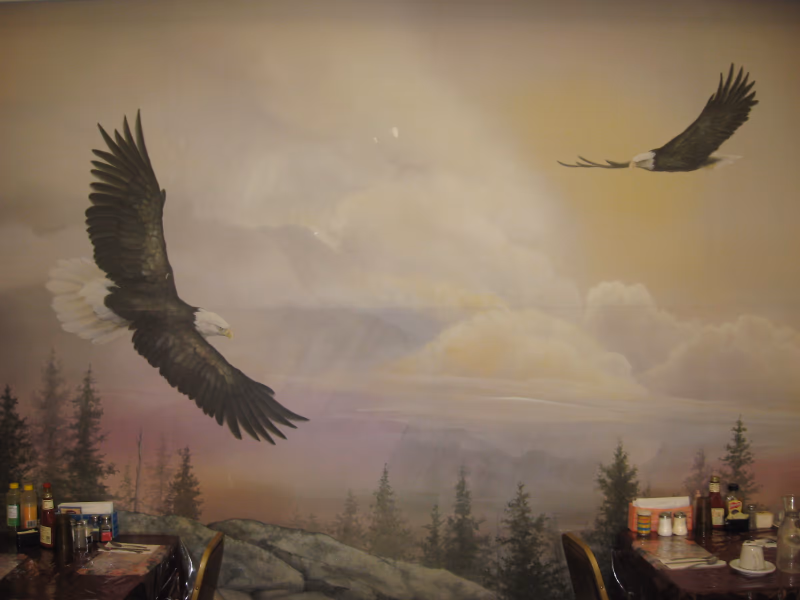 Interior view of a dining area with tables set with condiments, napkins, and cups. The wall features a large mural of two bald eagles flying over a forested landscape with clouds and a sunset sky.