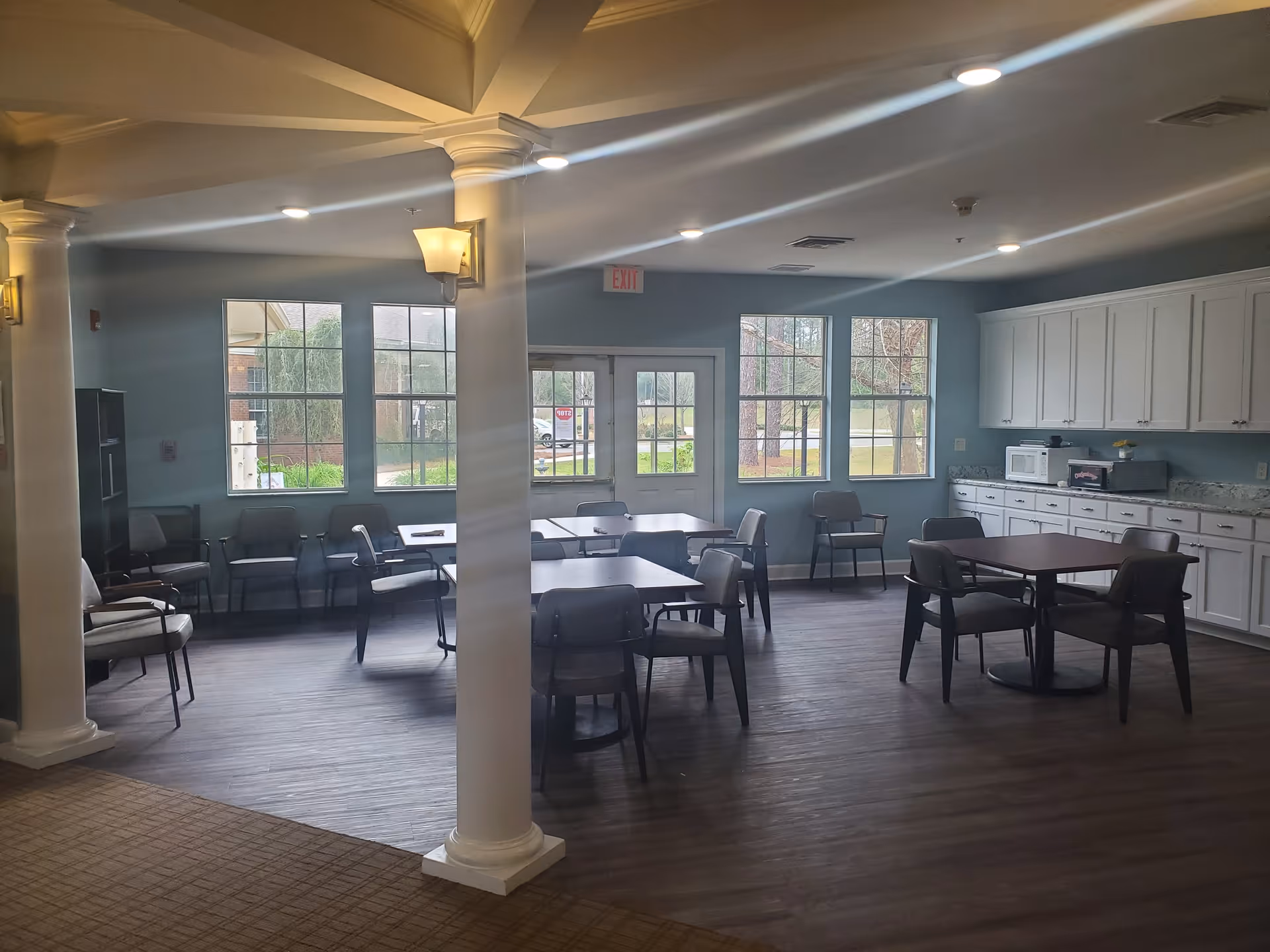 Interior view of a communal dining area with several tables and chairs arranged on a wooden floor. The room has large windows letting in natural light, white cabinets along one wall with a microwave and other appliances on the countertop, and decorative columns near the entrance.