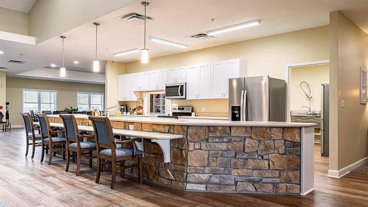 A spacious kitchen area in a senior living facility with a long stone-faced island counter and several wooden chairs with cushions lined up along it. The kitchen features white cabinets, a stainless steel refrigerator, a microwave, and an oven. Pendant lights hang from the ceiling above the counter, and the floor is covered with light wood flooring. In the background, there is a glimpse of another room with a sink and medical equipment.