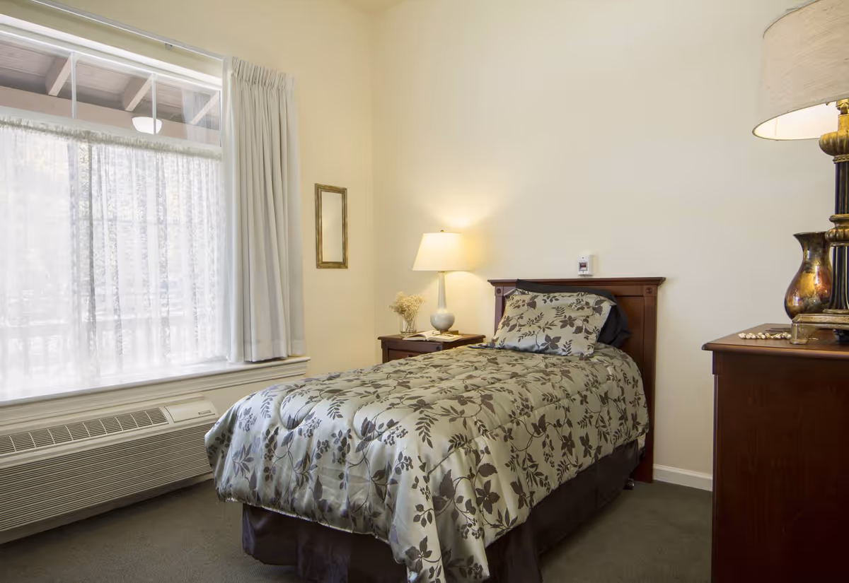 A small bedroom with a single bed covered in a floral patterned comforter. There is a wooden headboard, a nightstand with a lamp and dried flowers, a window with sheer and solid curtains, and a dresser with a lamp and decorative items.