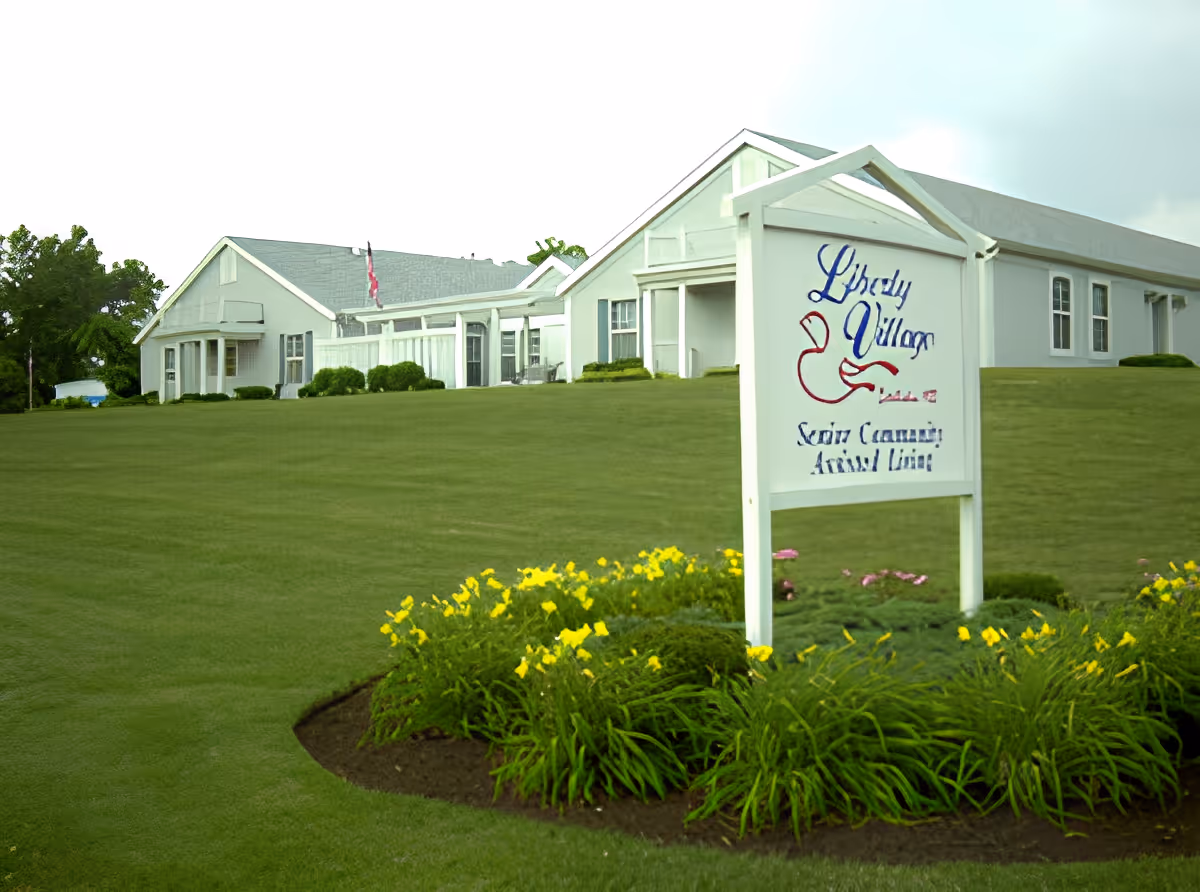 Exterior view of Liberty Village Senior Community building with a large green lawn and a flower bed with yellow flowers in front of a white sign displaying the facility's name.