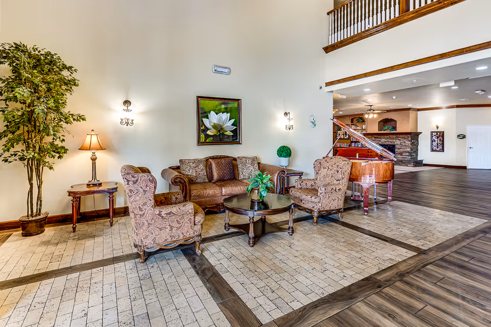 A spacious senior living facility common area with a seating arrangement including a brown sofa, two patterned armchairs, and a round wooden coffee table with a plant on it. There is a large potted plant and a side table with a lamp on the left. A grand piano is visible in the background near a stone fireplace. The room has high ceilings, light-colored walls, and a mix of tiled and wooden flooring.