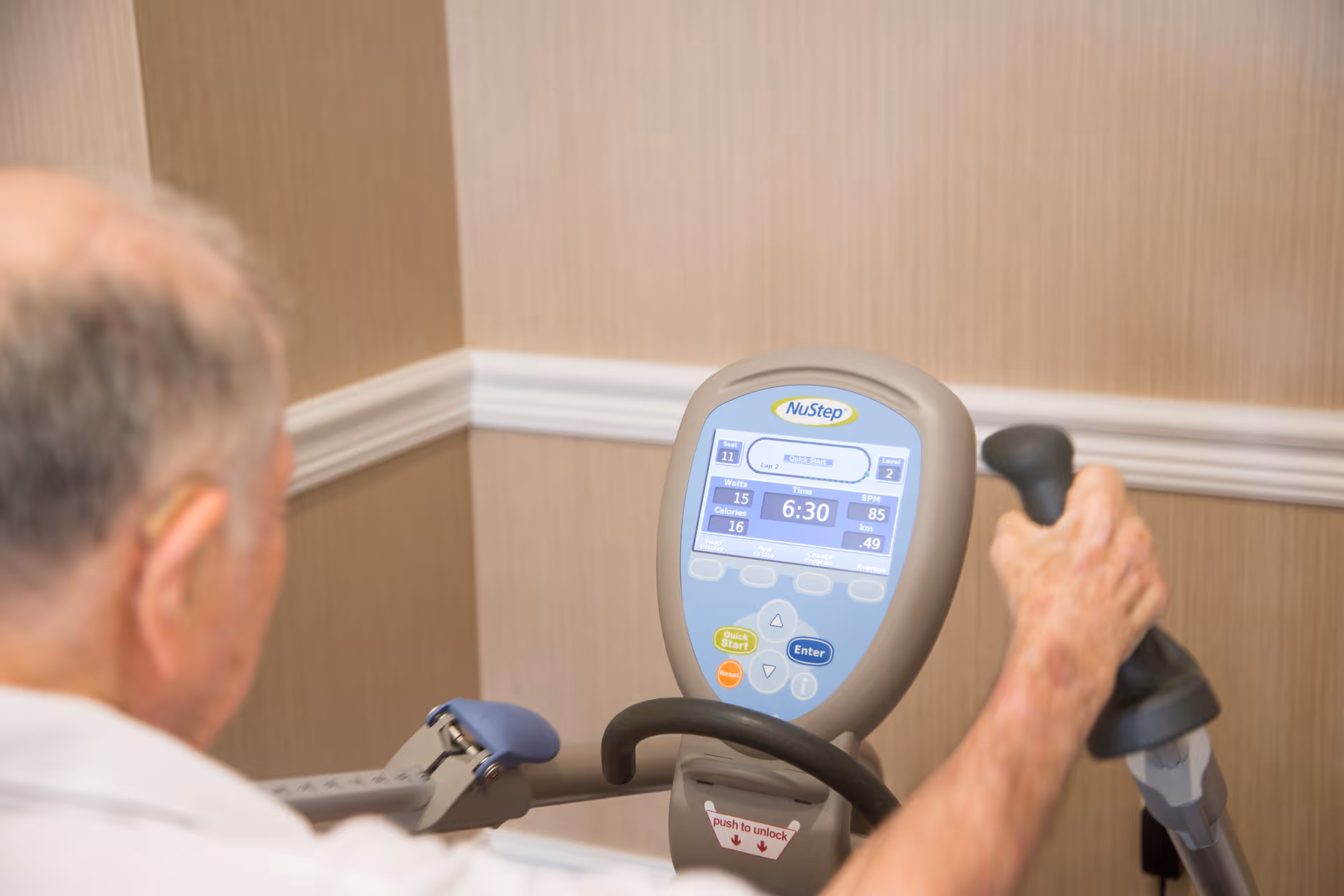 An elderly person using a NuStep exercise machine in a room with beige walls and white trim.