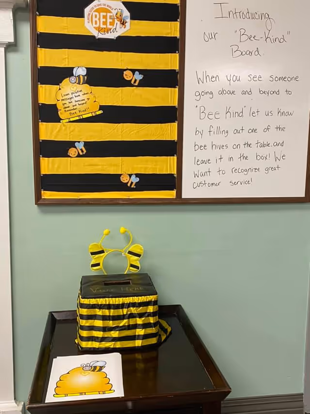 A bulletin board and a small table with a bee-themed kindness recognition setup. The bulletin board has black and yellow stripes with bee decorations and a note explaining the 'Bee-kind' board, encouraging people to recognize great customer service by filling out bee hive notes and leaving them in a decorated box. The table below holds the bee hive notes and a box covered in black and yellow stripes with bee antennae on top.