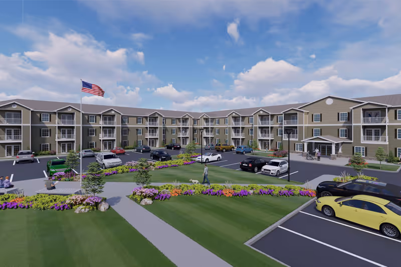 Exterior view of a three-story senior living community building with balconies, a parking lot with several cars, landscaped green areas with colorful flowers, a walking path, and an American flag on a flagpole under a partly cloudy sky.