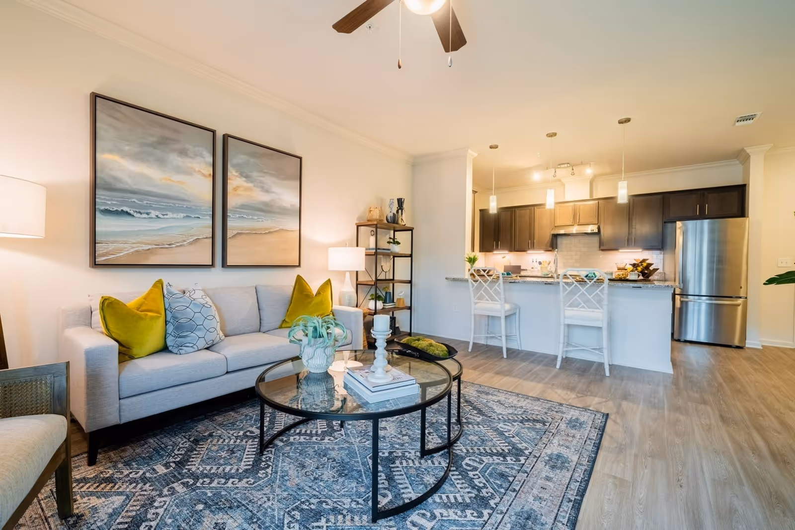 A modern living room and kitchen area with a light gray sofa adorned with green and patterned pillows, a round glass coffee table with decorative items, a patterned blue rug, and two large paintings of a beach scene on the wall. The kitchen features dark wood cabinets, a stainless steel refrigerator, a granite countertop with two white bar stools, and pendant lighting.