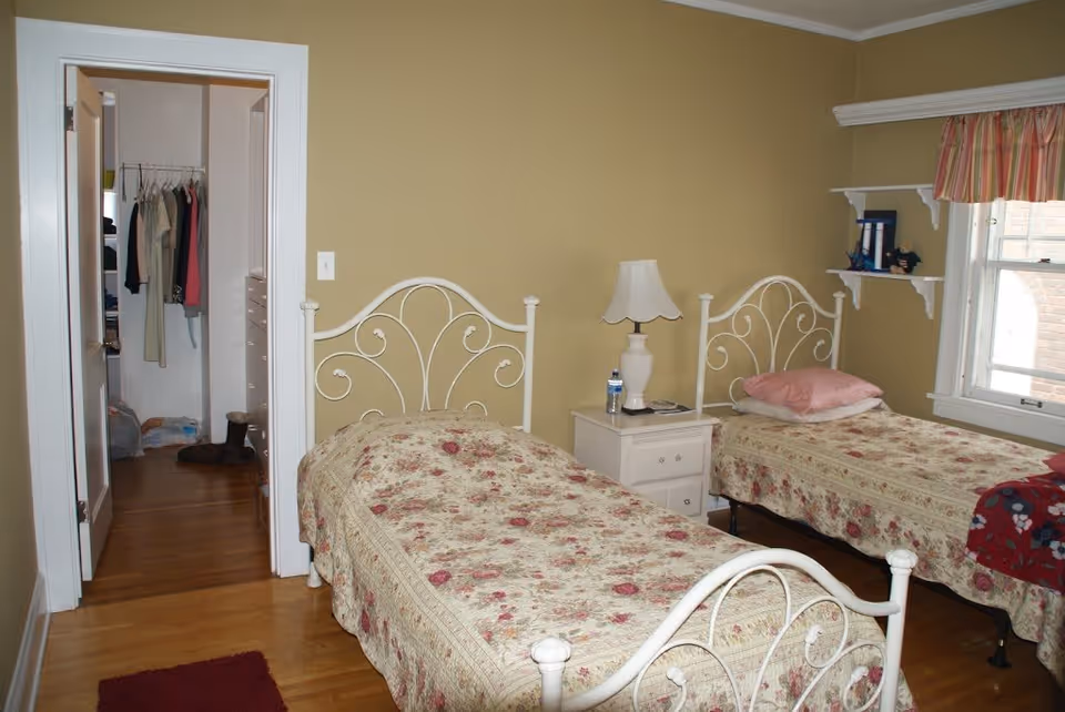 A bedroom with two twin beds featuring white metal headboards and floral bedspreads. Between the beds is a white nightstand with a lamp, a water bottle, and a phone. A window with a striped valance is on the right wall, and there are two white shelves with decorative items above the right bed. An open door on the left reveals a closet with hanging clothes and shelves.