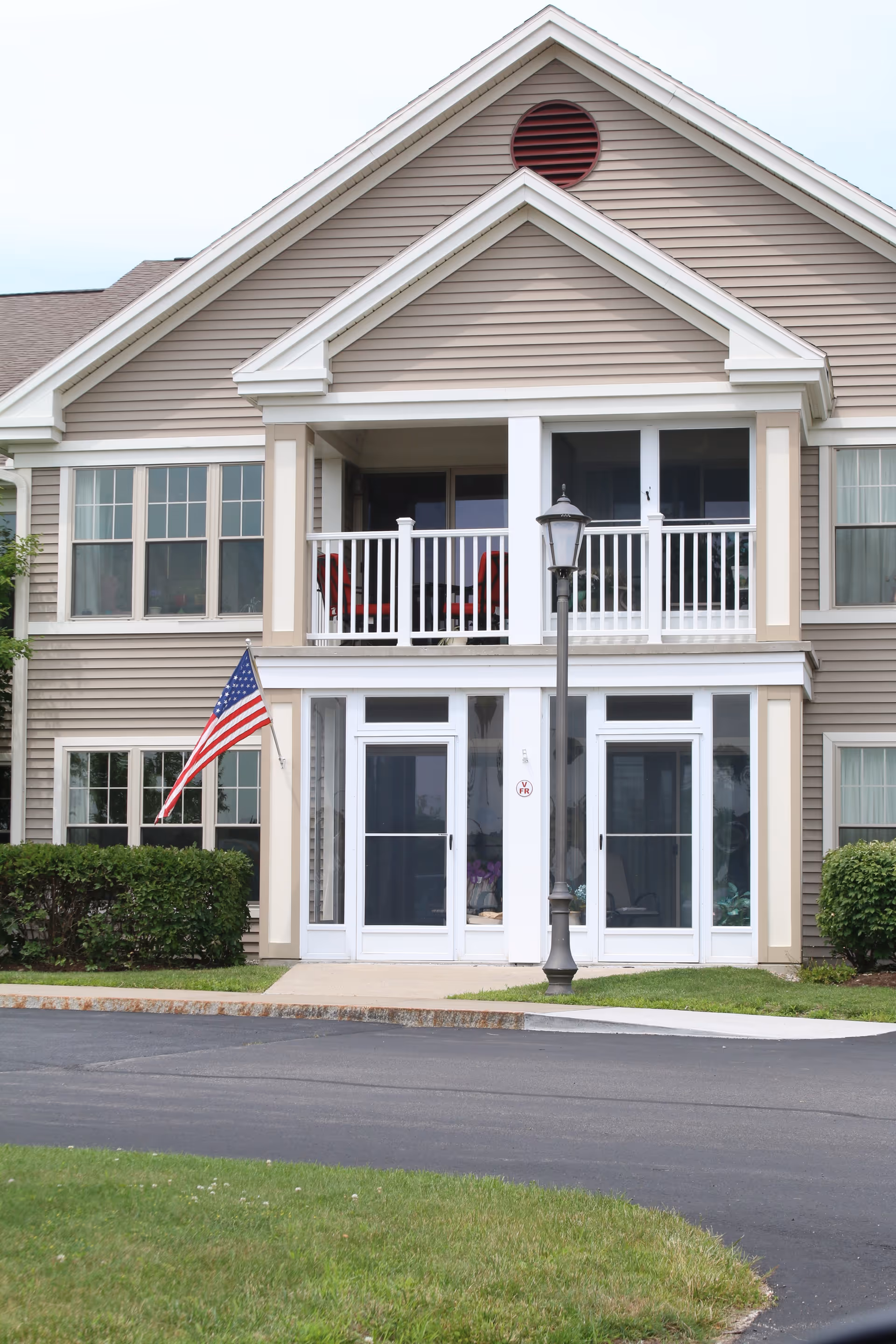 Exterior view of a two-story residential building with beige siding and white trim. The building features a balcony with white railing on the second floor and two glass doors on the ground floor. An American flag is displayed on the left side near some bushes, and a street lamp is positioned in front of the building. The area is landscaped with grass and a paved driveway.