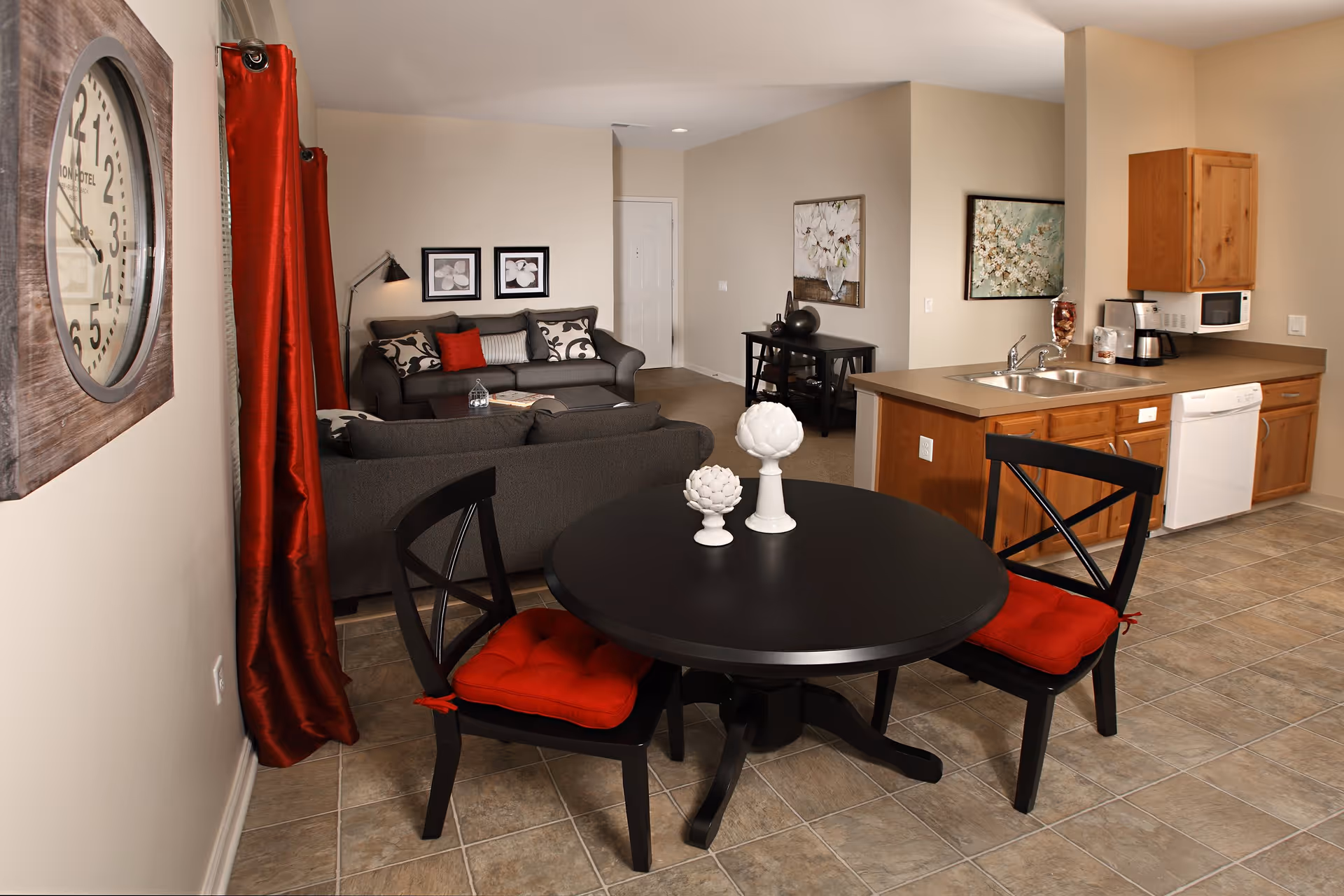 Interior view of a living space in a senior living facility named Avondale, featuring a small round black dining table with two black chairs with red cushions, a kitchen area with wooden cabinets, a sink, dishwasher, microwave, and coffee maker, and a living room area with two gray sofas, decorative pillows, wall art, and a large clock on the wall with red curtains beside it.
