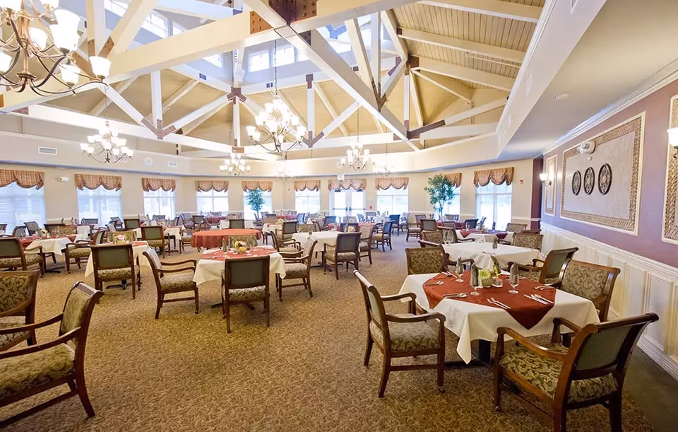 A spacious dining room with multiple round tables covered with white and red tablecloths, each set with silverware, glasses, and small floral centerpieces. The room features high vaulted ceilings with exposed wooden beams and several chandeliers providing warm lighting. Large windows with valances allow natural light to fill the space, and there are decorative wall panels and framed art on the walls.