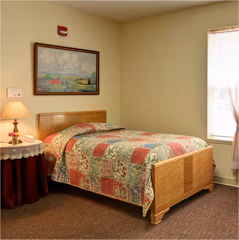 A cozy bedroom with a single wooden bed covered by a colorful patchwork quilt. Next to the bed is a round side table draped with a burgundy cloth and a lace overlay, holding a lamp and a small flower arrangement. A window with sheer curtains allows natural light into the room, and a framed landscape painting hangs on the wall above the bed.