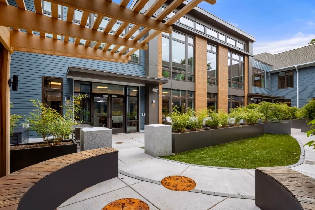 Outdoor courtyard area of a senior living facility with modern architecture, featuring a wooden pergola, curved wooden benches, large planters with green plants, and a well-maintained lawn. The building has large windows and a blue and wood-paneled exterior.