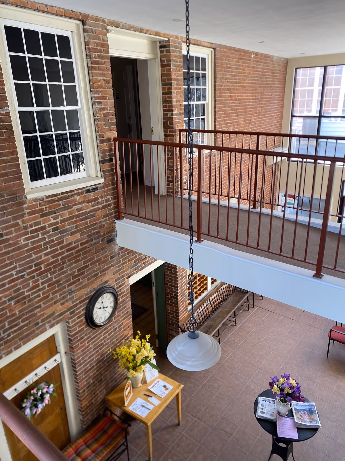 View from an upper floor overlooking a spacious interior area with brick walls and large windows. There is a hanging light fixture in the center, a wooden table with yellow flowers and papers, a round black table with purple flowers and magazines, and a bench along the far wall. A clock is mounted on the brick wall near a wooden door decorated with a small wreath.