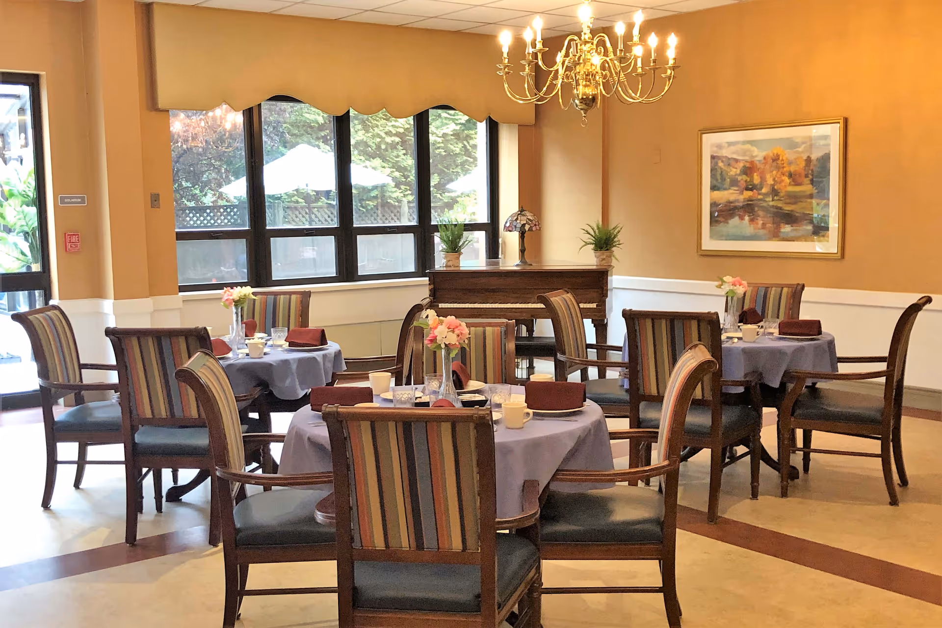 A dining room with round tables covered in light purple tablecloths, each set with cups, glasses, plates, and maroon napkins. The chairs have wooden frames with striped upholstery. A chandelier hangs from the ceiling, and a piano with two potted plants and a decorative lamp sits against the wall. Large windows show an outdoor area with umbrellas and greenery. A framed painting of a landscape is on the wall.