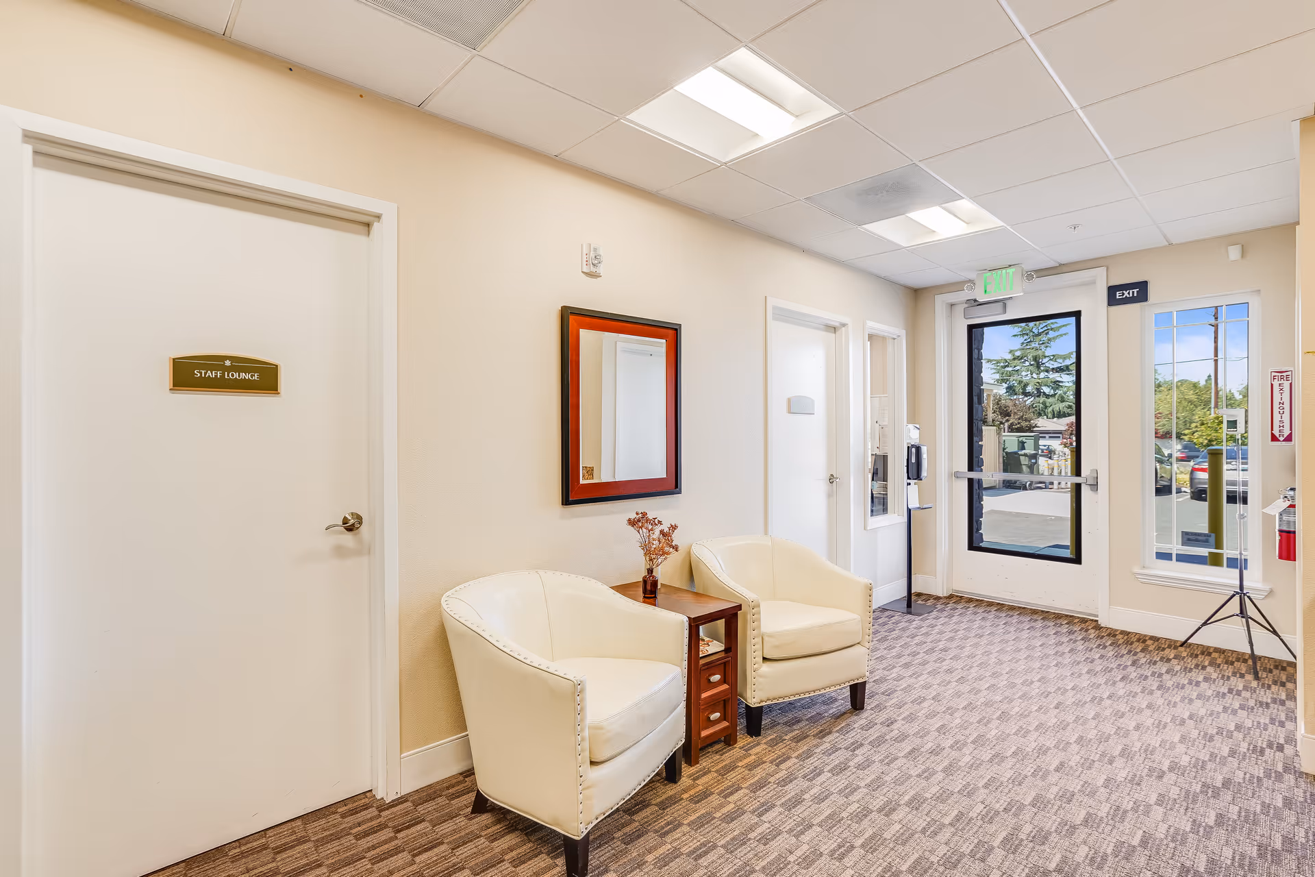 A hallway area in a senior living facility with two cream-colored armchairs and a small wooden table with a vase of dried flowers between them. There is a mirror with a red and black frame on the wall above the chairs. To the left is a door labeled 'Staff Lounge.' At the end of the hallway is a glass exit door with an illuminated green exit sign above it, and a window to the right showing a parking lot and trees outside.
