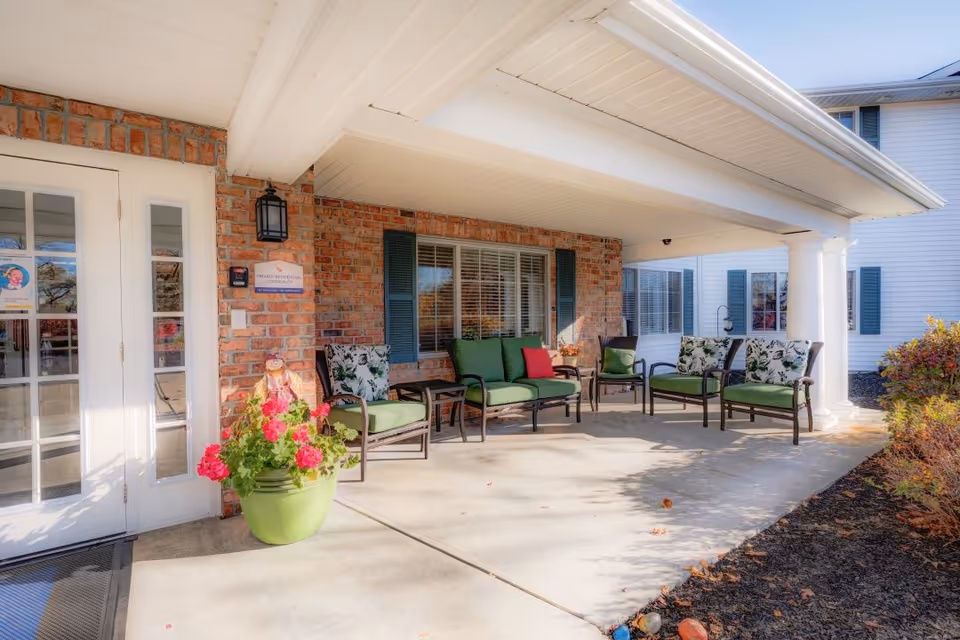 Covered outdoor seating area at the entrance of a building with brick walls and white pillars. The seating includes green cushioned chairs and a loveseat with floral pillows, arranged around small tables. There is a large green planter with pink flowers near the white double doors, and some bushes and landscaping along the walkway.