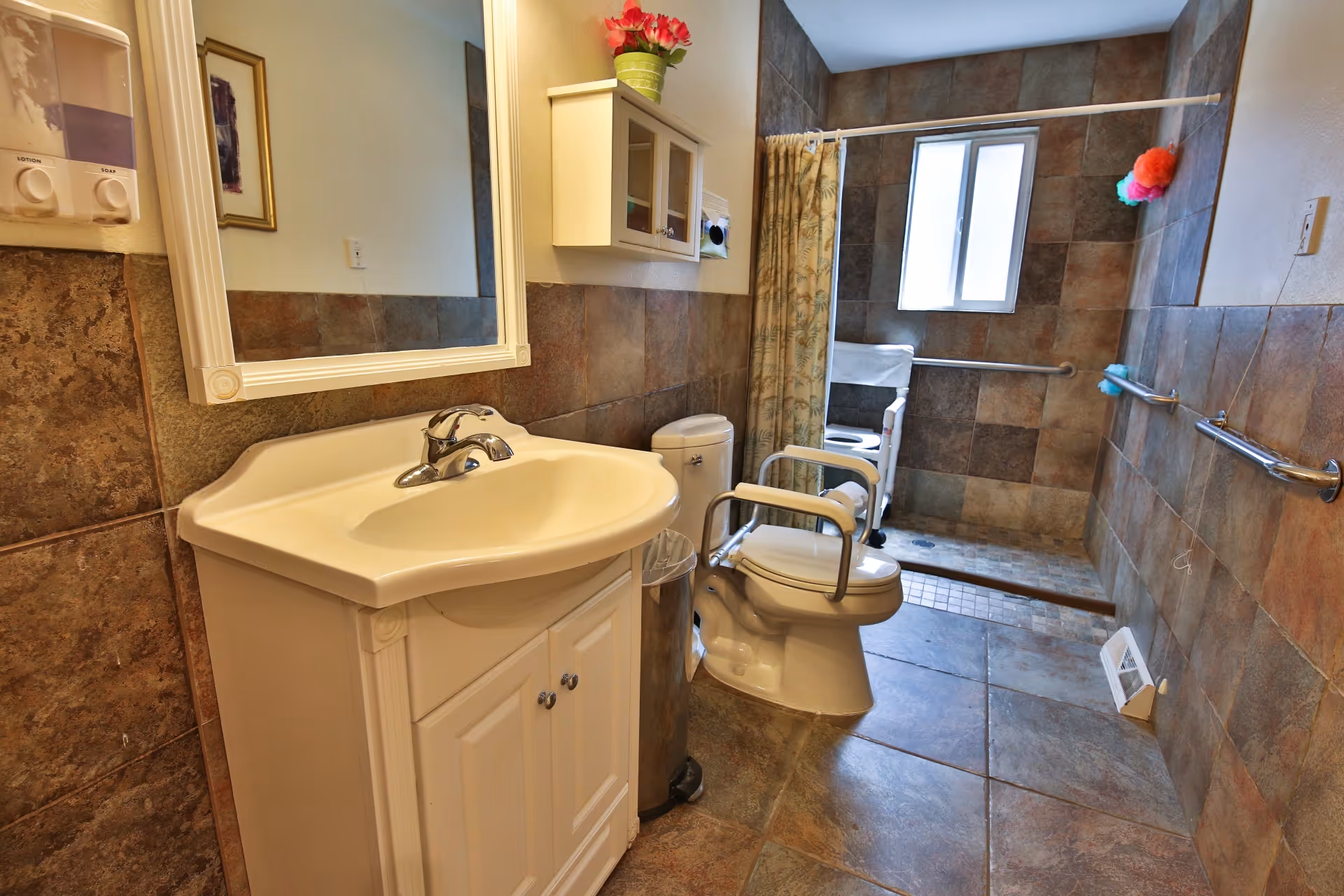 A bathroom with brown tiled walls and floor, featuring a white sink with a mirror above it, a toilet with armrests, and a walk-in shower area with a window and grab bars. There is a small cabinet mounted on the wall and a soap and lotion dispenser near the mirror.