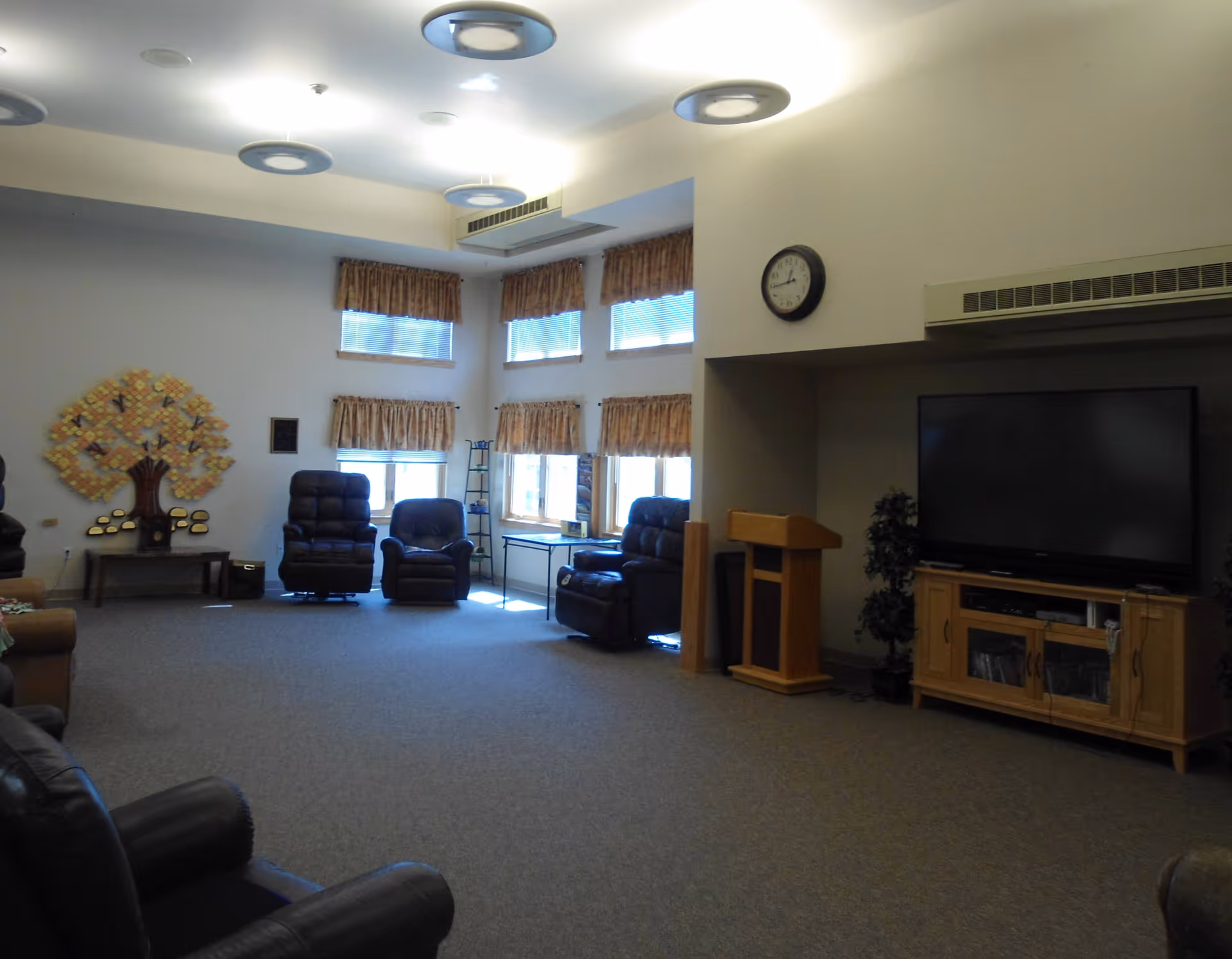 A spacious senior living common room with leather recliners, a large TV on a wooden cabinet, a podium, a wall clock, and windows with valances.