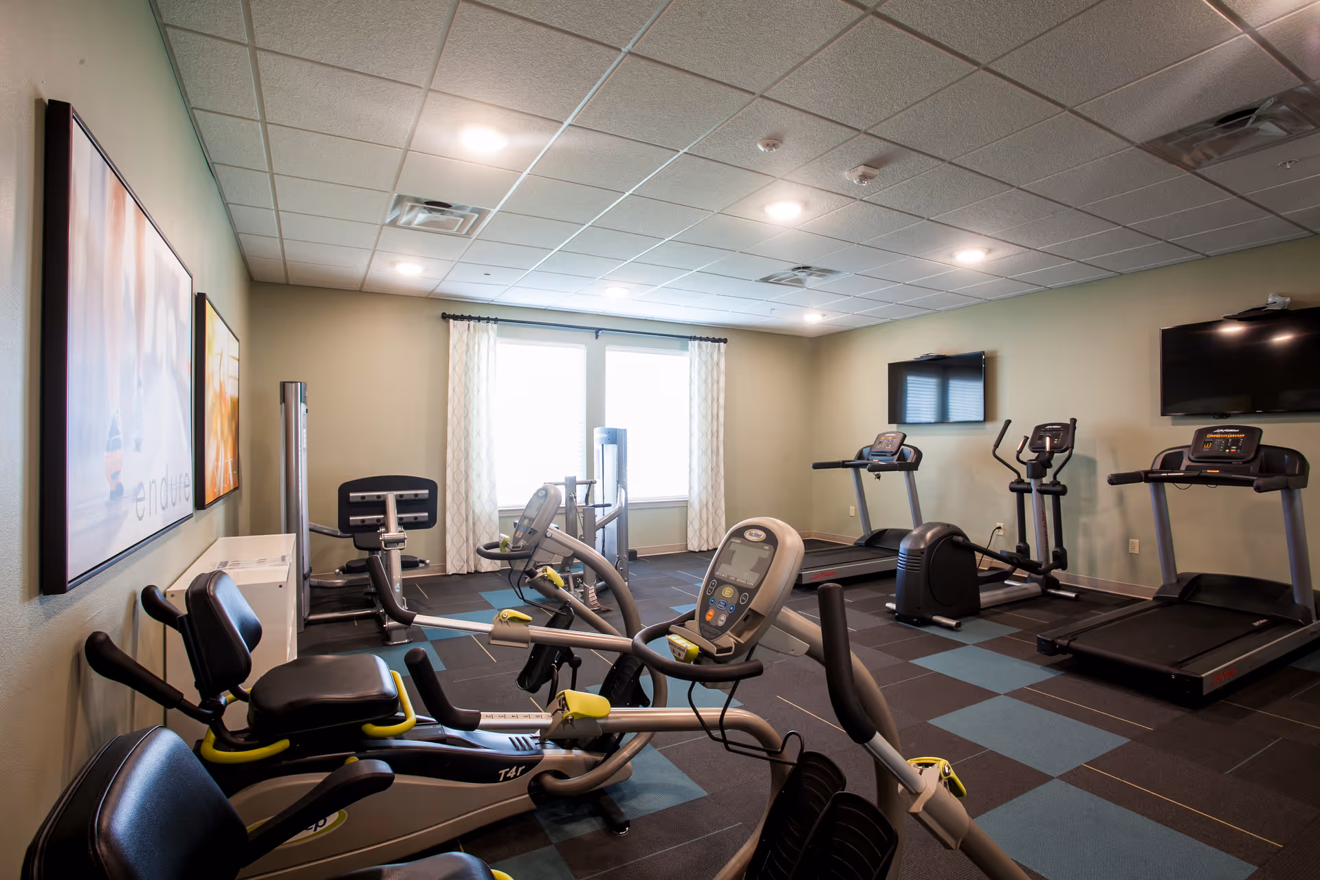 Interior view of a fitness room with exercise equipment including treadmills, stationary bikes, and weight machines. The room has a tiled ceiling with recessed lighting, a window with curtains, two wall-mounted TVs, and framed artwork on the wall.