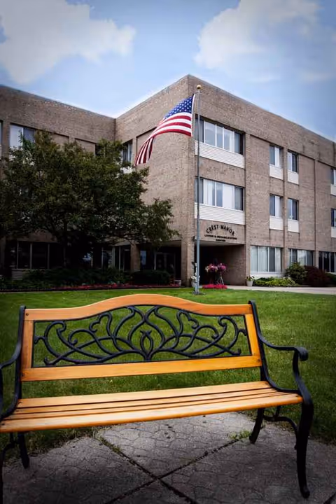 Front exterior of Crest Manor Living & Rehab Center with flagpole and bench.