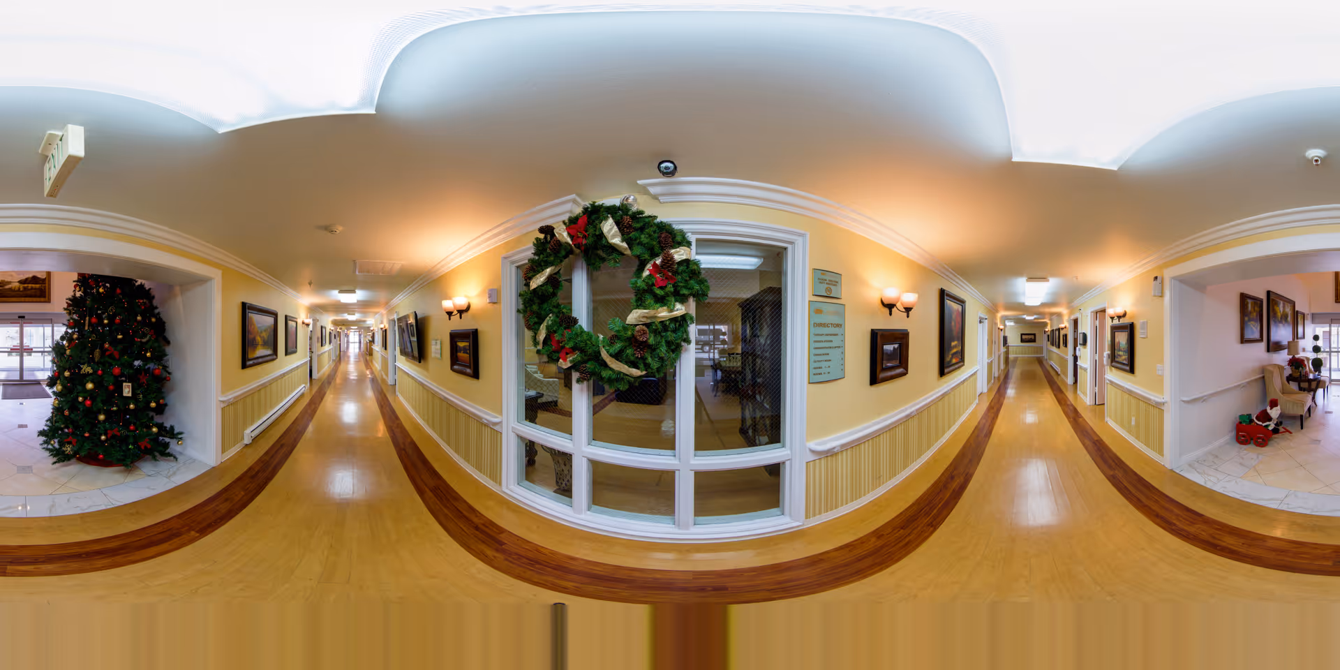 A long, well-lit hallway in a senior living facility decorated for the holidays with a large Christmas tree on the left side and a festive wreath hanging on a window in the center. The hallway has wooden flooring, yellow walls with white trim, framed pictures on the walls, and wall-mounted light fixtures. There are doorways and windows along the hallway, and a small seating area with holiday decorations is visible on the right.