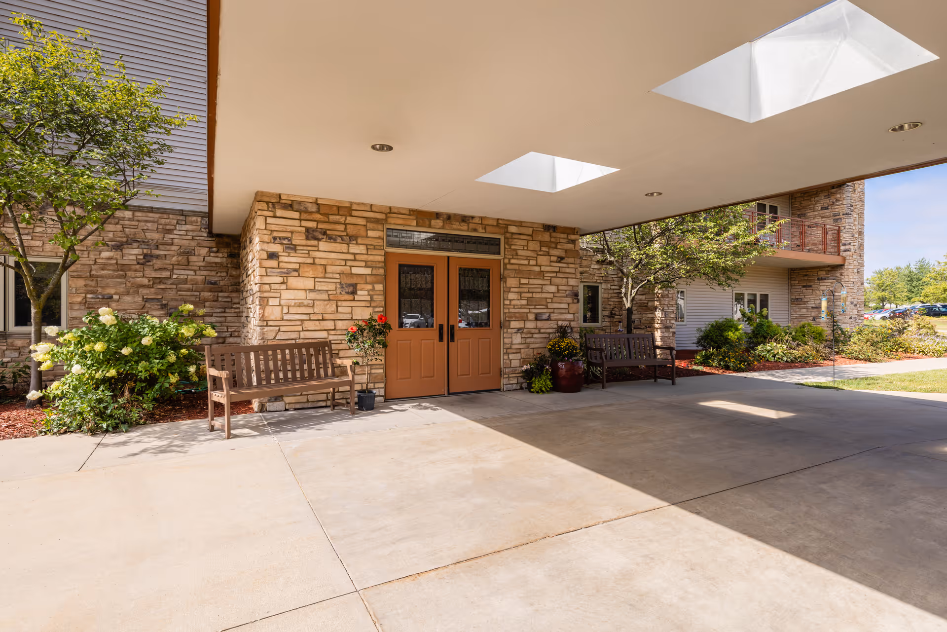Entrance area of a senior living facility with double wooden doors set in a stone wall. There are two wooden benches on either side of the doors, potted plants, and small trees with green foliage. The area is covered by a roof with skylights, and there is a concrete driveway in front.