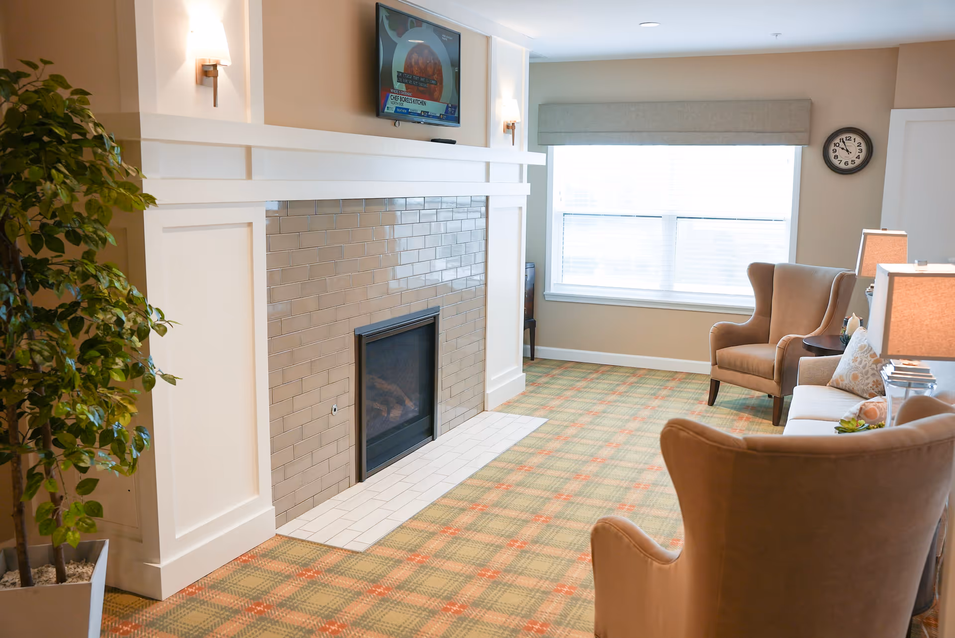 A cozy living room area with a fireplace featuring beige tiles and a white mantel. Above the fireplace is a mounted flat-screen TV. The room has a large window with blinds and a beige valance, a wall clock, and comfortable seating including two beige armchairs and a sofa with decorative pillows. There are two table lamps on side tables and a potted plant near the fireplace. The carpet has a plaid pattern in green and orange tones.