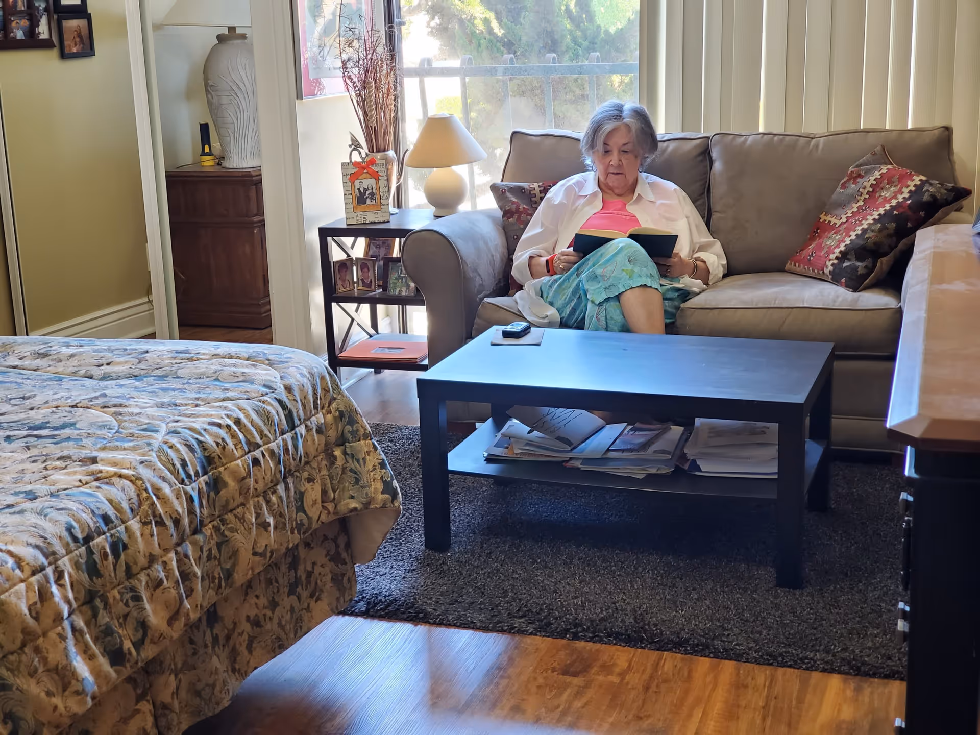 An elderly woman sitting on a beige couch in a cozy living area, reading a book. The room features a patterned bedspread on a bed to the left, a black coffee table with magazines and papers underneath, a side table with framed photos and a lamp, and vertical blinds covering a window behind the couch.