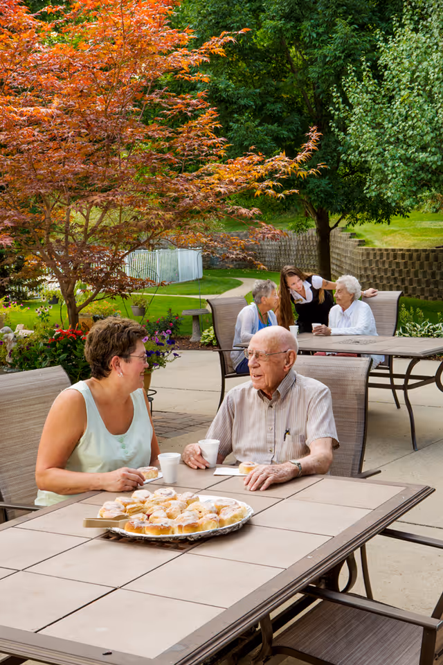 An outdoor patio area at Village Ridge Assisted Living with elderly residents and a caregiver sitting and chatting around tables. There are trees with green and orange leaves, flowers, and a paved walkway in the background. A tray of pastries and cups are on the table in the foreground.