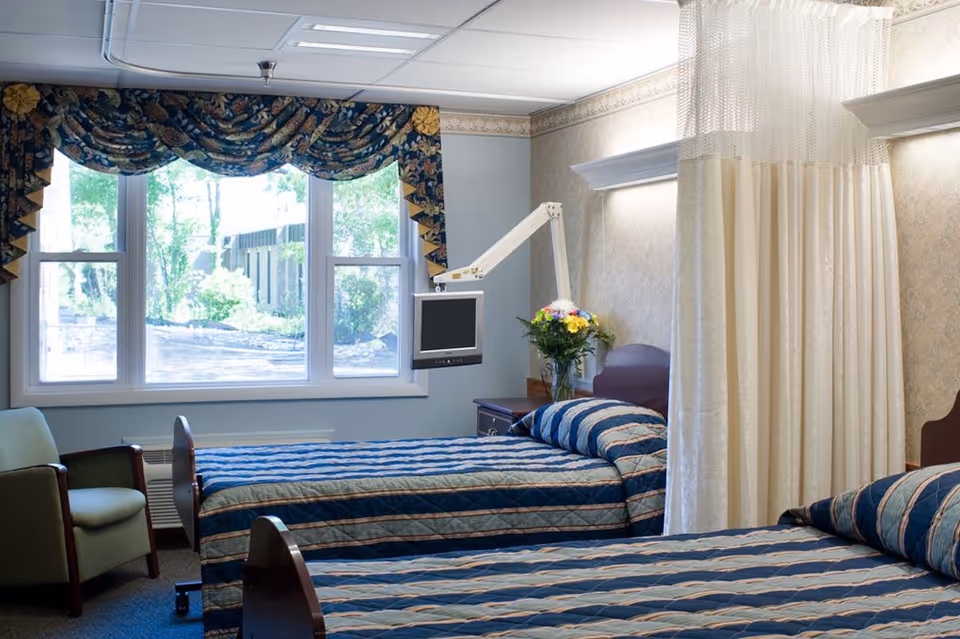 A room with two single beds covered in blue and beige striped bedspreads, a green armchair near a large window with floral valance curtains, a wall-mounted TV on an adjustable arm, a vase of flowers on a nightstand, and a privacy curtain partially drawn between the beds.