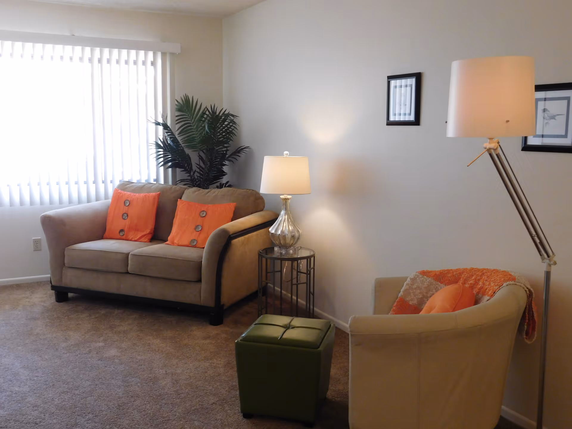 A cozy living room with a beige loveseat adorned with two orange pillows, a beige armchair with an orange pillow and throw blanket, a green ottoman, a side table with a lamp, a tall floor lamp, two framed pictures on the wall, and a large window with vertical blinds letting in natural light.